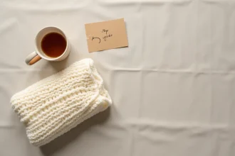Cadeaux de naissance minimalistes disposés en flat lay sur lin brut : couverture en laine crème, tasse en céramique et carte kraft manuscrite